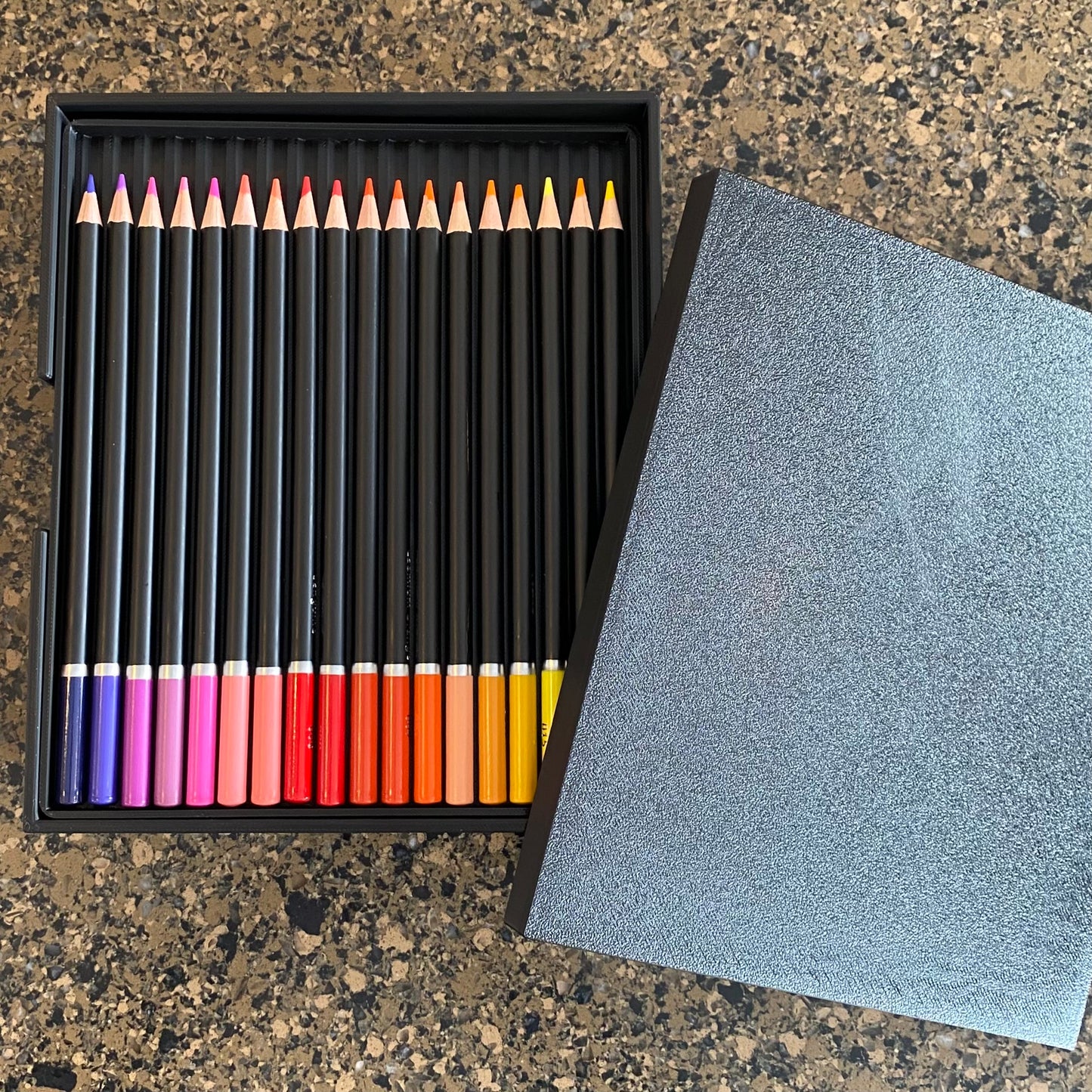 A black colored pencil caddy with multiple colored pencils visible, placed next to a gray lid or cover.