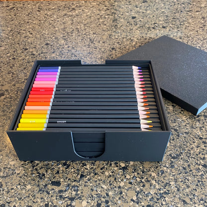 A black colored pencil storage caddy open on a surface displaying multiple colored pencils inside, with the caddy lid placed beside it.
